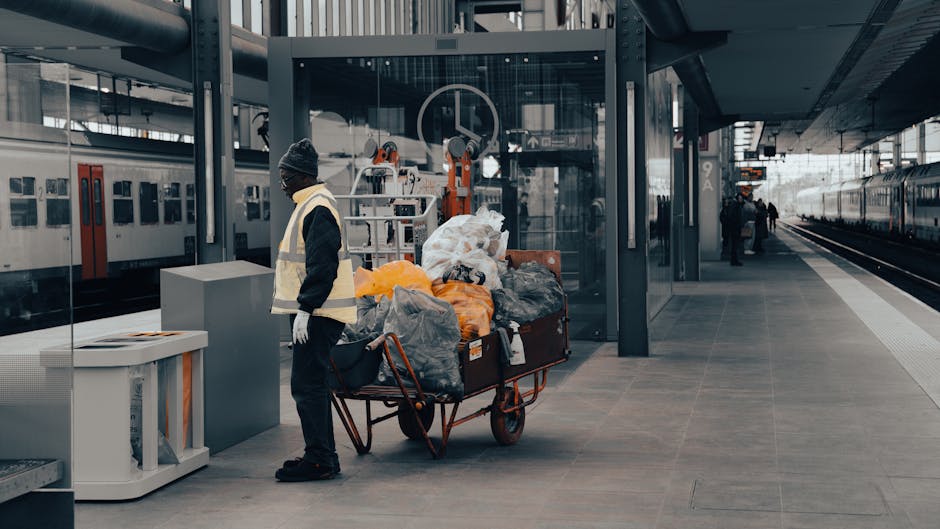 A narrow urban alleyway filled with a large accumulation of mixed waste and rubbish. In the foreground, there is a metal hand truck or trolley loaded with black and grey refuse bags, some appearing sl